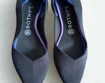 May include: A pair of dark gray pointed-toe flats. The shoes have a textured knit upper with a blue trim around the opening and a purple accent along the inner edge. The brand name "ROTHY'S" is printed on the insole.