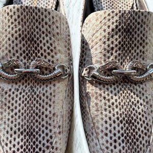 May include: A pair of slip-on shoes with a snakeskin pattern in shades of beige and brown. Each shoe features a silver-colored chain detail with dragon head accents. The shoes have a pointed toe and a low heel.