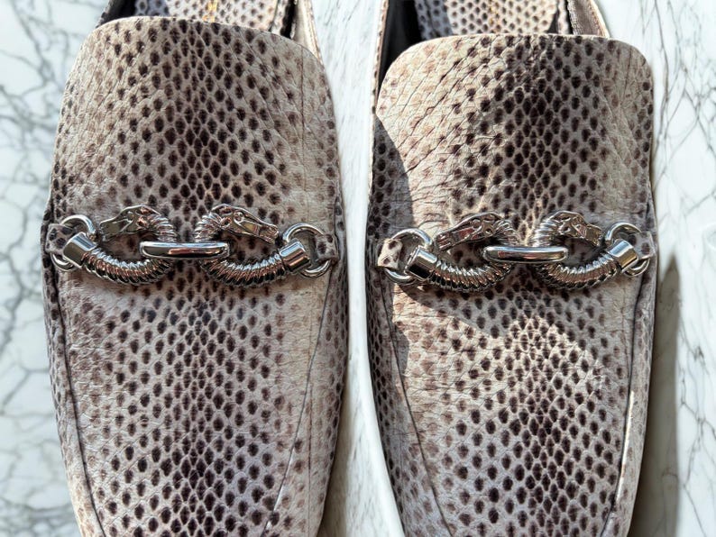 May include: A pair of slip-on shoes with a snakeskin pattern in shades of beige and brown. Each shoe features a silver-colored chain detail with dragon head accents. The shoes have a pointed toe and a low heel.