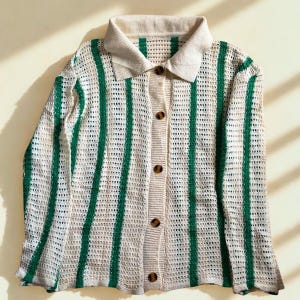 May include: A cream-colored, open-weave cardigan with vertical green stripes. The collared shirt has a button-down front with brown buttons. The collar and cuffs are a solid cream color. The cardigan is a lightweight knit.