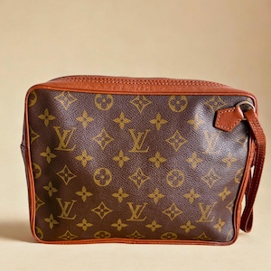 Puede incluir: Un bolso marrón y beige con un patrón de logotipo repetido. El bolso tiene forma rectangular con cierre de cremallera y una correa de muñeca de cuero. El ribete y la correa son de color marrón oscuro.