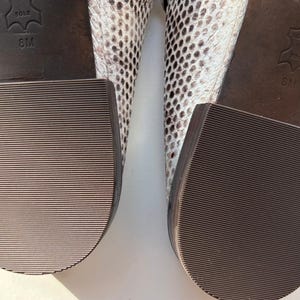 May include: Close-up of the soles of two shoes. The soles are brown with a ridged texture. The heels are thick and square. The uppers of the shoes are patterned with a snakeskin design in shades of white, gray, and black. The size is 8M.