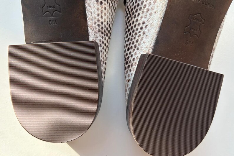 May include: Close-up of the soles of two shoes. The soles are brown with a ridged texture. The heels are thick and square. The uppers of the shoes are patterned with a snakeskin design in shades of white, gray, and black. The size is 8M.