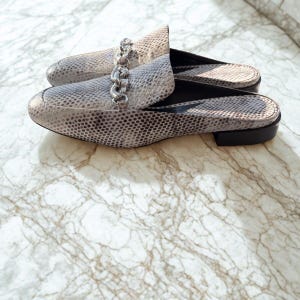 May include: A pair of gray and white snakeskin-patterned mules with a silver chain detail across the top. The shoes have a low heel and are displayed on a marble surface.