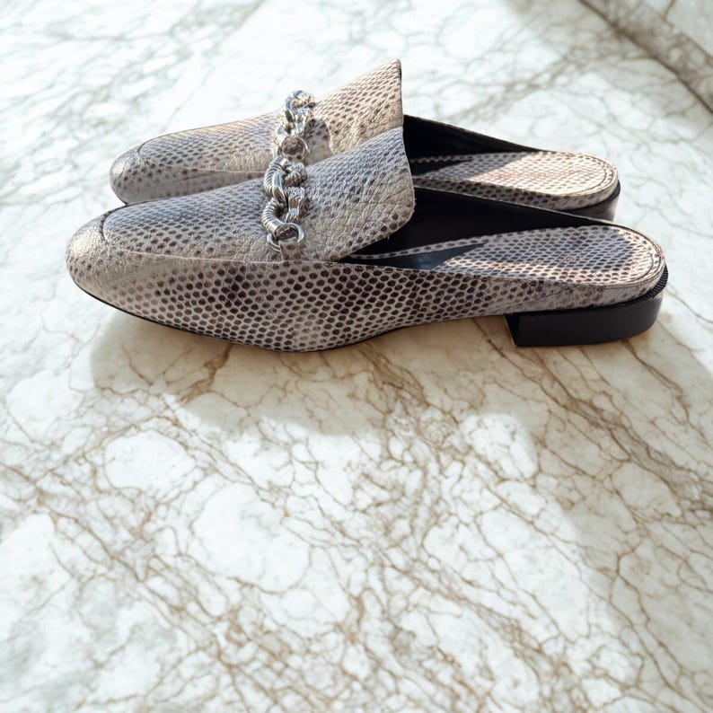 May include: A pair of gray and white snakeskin-patterned mules with a silver chain detail across the top. The shoes have a low heel and are displayed on a marble surface.