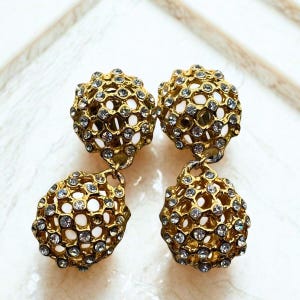 May include: Four gold-toned earrings with a spherical design, encrusted with small, clear crystals. The earrings have a hollow, openwork structure, with a smaller, teardrop-shaped element hanging below the main sphere. The earrings are displayed on a white surface.