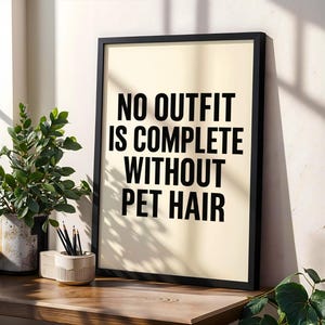 Op de afbeelding: Een ingelijste print met de tekst "NO OUTFIT IS COMPLETE WITHOUT PET HAIR" in zwarte letters. De print zit in een zwarte lijst en wordt weergegeven op een houten oppervlak. Een plant in een pot en een potloodhouder zijn ook aanwezig.
