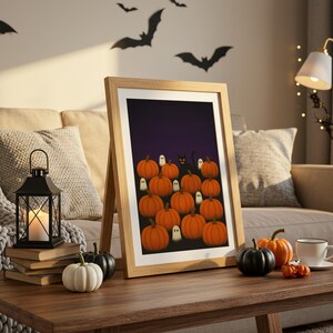 May include: A framed Halloween-themed print featuring orange pumpkins, white ghosts, and a black cat against a dark purple background. The print is displayed on a wooden table with decorative pumpkins and a lit candle lantern.