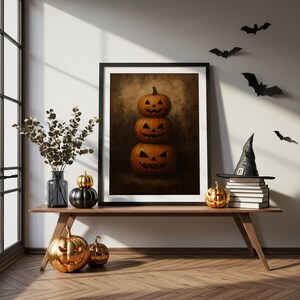 May include: A framed Halloween art print featuring three carved pumpkins stacked on top of each other. The scene is decorated with a witch's hat, bats, and metallic pumpkin decor on a wooden table. The pumpkins have carved faces.
