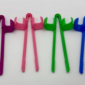 May include: A collection of colorful plastic tongs in black, purple, pink, green, blue, and orange. Each tong has a long handle and a curved head with a fork-like design, suitable for various kitchen tasks.