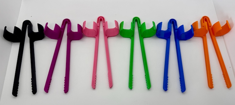 May include: A collection of colorful plastic tongs in black, purple, pink, green, blue, and orange. Each tong has a long handle and a curved head with a fork-like design, suitable for various kitchen tasks.