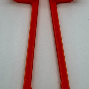 May include: Bright orange plastic tool with two long, parallel arms connected at the top by a curved piece. The ends of the arms have a scalloped edge. The tool is designed to hold or grip something.