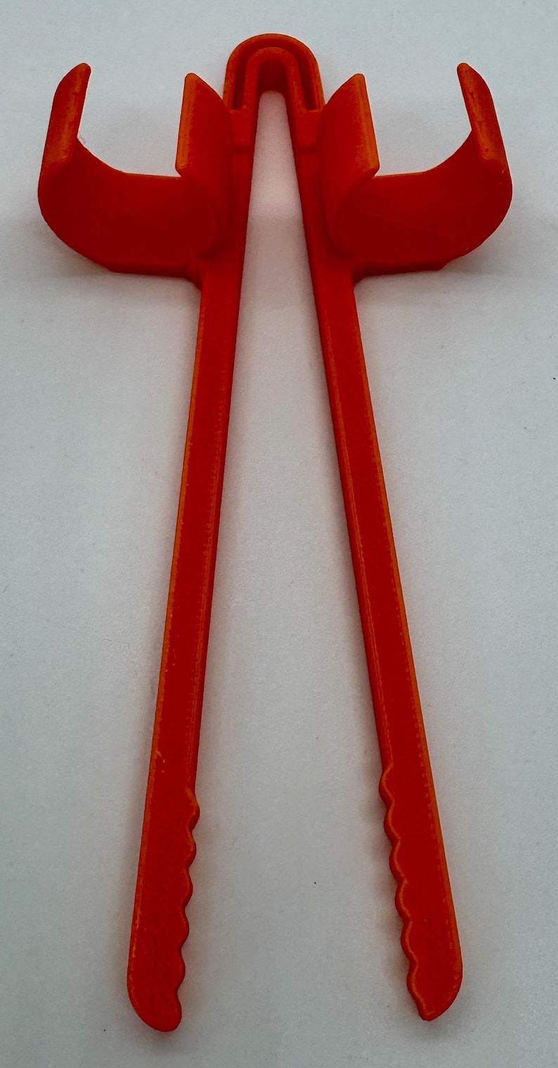 May include: Bright orange plastic tool with two long, parallel arms connected at the top by a curved piece. The ends of the arms have a scalloped edge. The tool is designed to hold or grip something.