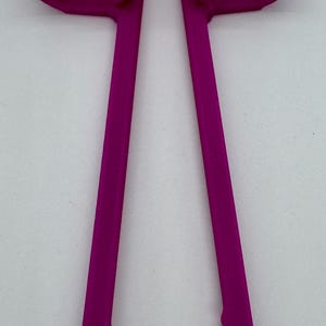 May include: A magenta-colored, plastic tool with a unique design. It features two curved arms connected by a bridge, with long, straight handles ending in a scalloped edge. The object is set against a plain white background.