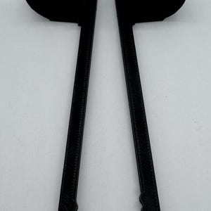 May include: A black, 3D-printed tool with two long, textured arms and curved ends. The tool has a central loop and is designed to hold or manipulate objects. The ends of the arms have a serrated design.