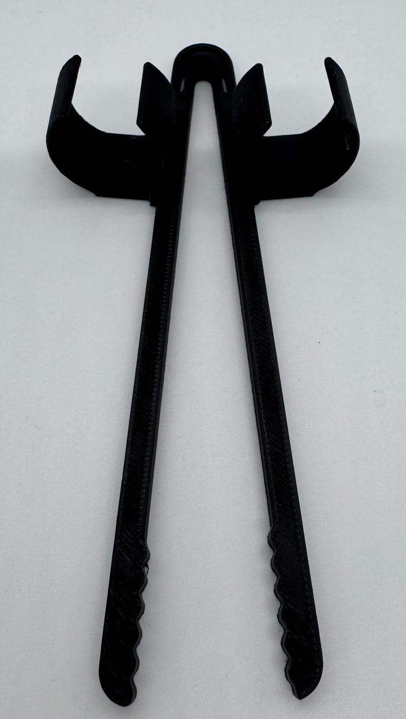 May include: A black, 3D-printed tool with two long, textured arms and curved ends. The tool has a central loop and is designed to hold or manipulate objects. The ends of the arms have a serrated design.