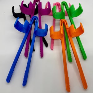 May include: A collection of colorful, plastic, long-handled reacher grabber tools in various colors, including blue, orange, green, pink, purple, and black. The tools have a curved handle and a claw-like grip at the end, designed for reaching and grabbing objects.