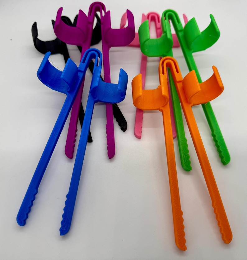 May include: A collection of colorful, plastic, long-handled reacher grabber tools in various colors, including blue, orange, green, pink, purple, and black. The tools have a curved handle and a claw-like grip at the end, designed for reaching and grabbing objects.