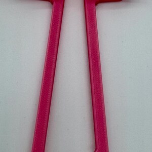May include: A bright pink, 3D-printed object with a U-shaped top and two curved arms. Two long, parallel legs extend downward, each with a scalloped edge. The object appears to be a tool or accessory.