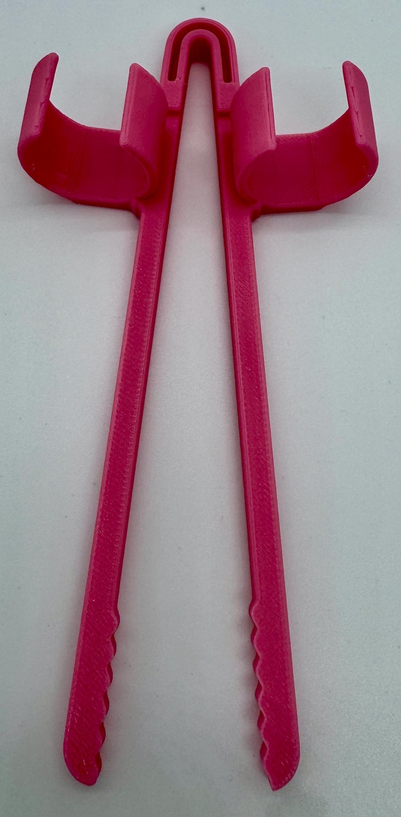 May include: A bright pink, 3D-printed object with a U-shaped top and two curved arms. Two long, parallel legs extend downward, each with a scalloped edge. The object appears to be a tool or accessory.