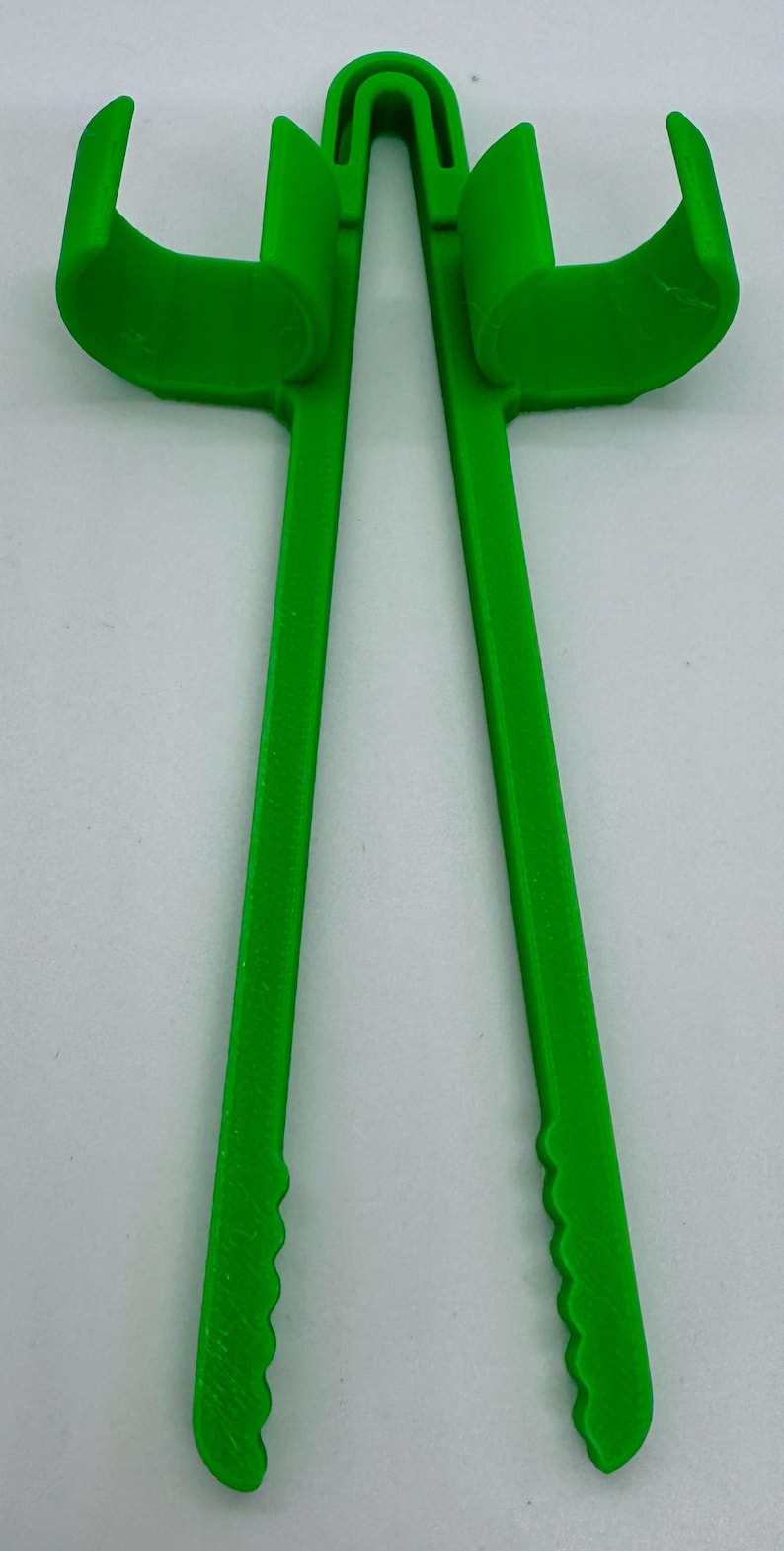 May include: Bright green 3D-printed tool with two curved arms and two long, parallel handles. The handles have a textured, scalloped edge. The tool is designed to hold or grip something.