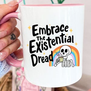 May include: White ceramic mug with a pink interior and handle. The mug features the text "Embrace the Existential Dread" with a cartoon skeleton drinking from a mug, set against a rainbow. The background includes pink flowers in a vase.