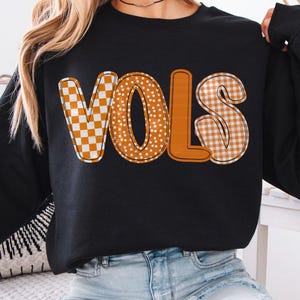 May include: Black long-sleeve sweatshirt with the word "VOLS" in orange and white checkered and polka dot patterns. The text "VOLS" is a collegiate design. The model is wearing a size M for a relaxed fit.