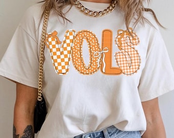 May include: Cream-colored t-shirt with the word "VOLS" in orange. The letters have different patterns: checkered, polka dots, and gingham. The shirt is paired with blue jeans.