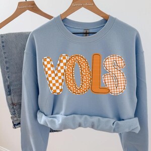 May include: A light blue sweatshirt with the word "VOLS" in orange letters. The letters have a checkerboard and polka dot pattern. The sweatshirt is hanging on a wooden hanger with a pair of blue jeans.