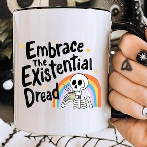 Könnte beinhalten: Weiße Keramik-Tasse mit schwarzem Henkel und Rand. Die Tasse trägt den Text "Embrace the Existential Dread" mit einem Skelett, das aus einer Tasse trinkt, vor einem Regenbogen. Die Tasse wird von einer Hand mit schwarz-weißer Nagelkunst gehalten.