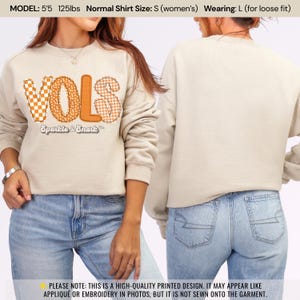 May include: Beige sweatshirt with the word "VOLS" in orange, with a checkerboard and dotted pattern. The text "Sparkle & Snark" is below the word "VOLS". The model is wearing a size L for a loose fit.