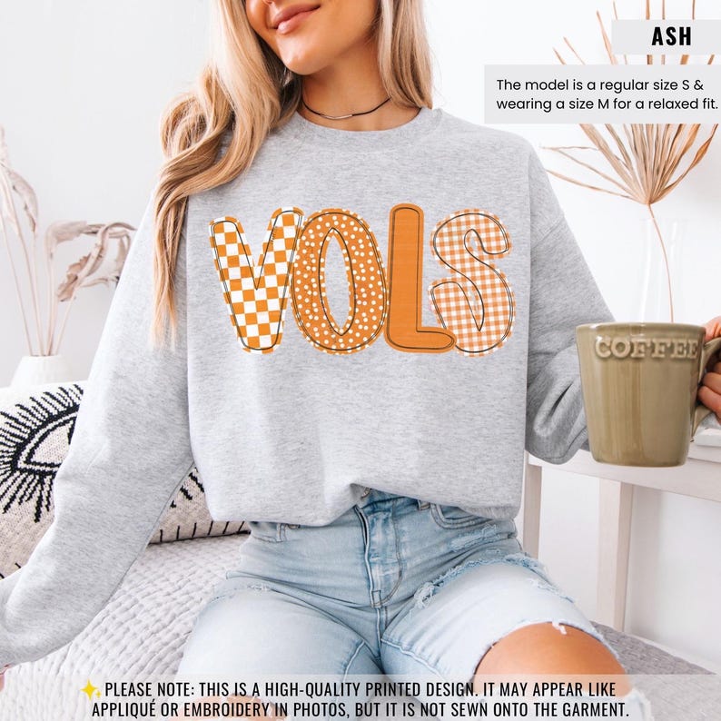 May include: Ash-colored sweatshirt with the word "VOLS" in orange and white patterned letters. The model is wearing a size M for a relaxed fit. A coffee mug is held in the hand. The text on the garment is a high-quality printed design.
