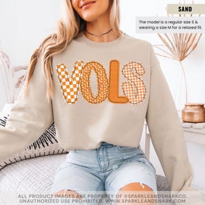 May include: A sand-colored sweatshirt with the word "VOLS" in orange, featuring a mix of patterns: a checkered "V", dotted "O", and a gingham "S". The model is wearing a size M for a relaxed fit. The text "SAND" is in the upper right corner.