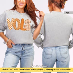May include: A light gray sweatshirt with the word "VOLS" in orange and checkered patterns. The sweatshirt is shown from the front and back, with the word "ASH" visible on the back. The model is wearing an XL for an oversized fit.