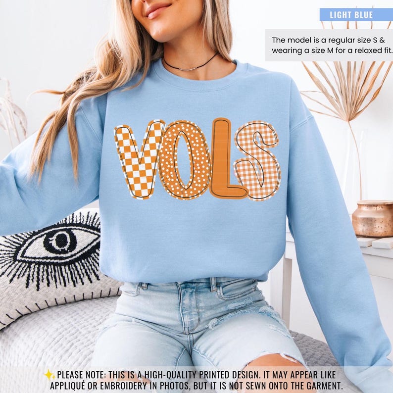 May include: Light blue sweatshirt with the word "VOLS" in orange. The letters have a mix of patterns, including checkerboard and polka dots. The text "The model is a regular size S & wearing a size M for a relaxed fit." is in the top right corner.