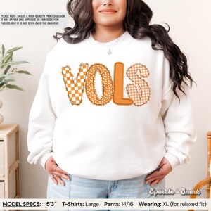 May include: White sweatshirt with the word "VOLS" in orange and patterned letters. The "V" is a checkered pattern, the "O" has a polka dot design, and the "S" is a gingham pattern. The text "Sparkle & Snark" is in the bottom right corner.