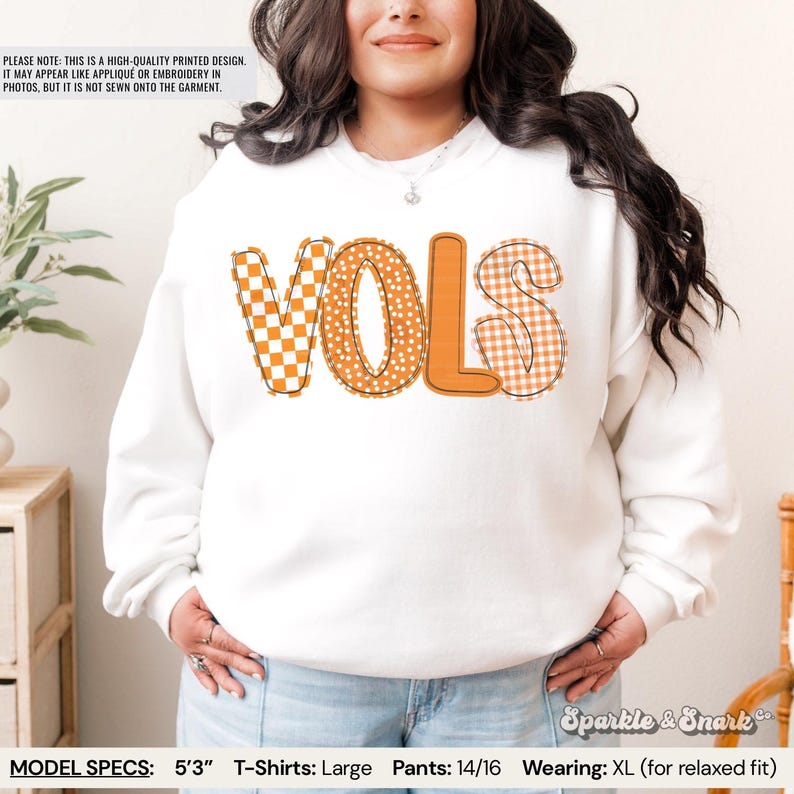 May include: White sweatshirt with the word "VOLS" in orange and patterned letters. The "V" is a checkered pattern, the "O" has a polka dot design, and the "S" is a gingham pattern. The text "Sparkle & Snark" is in the bottom right corner.