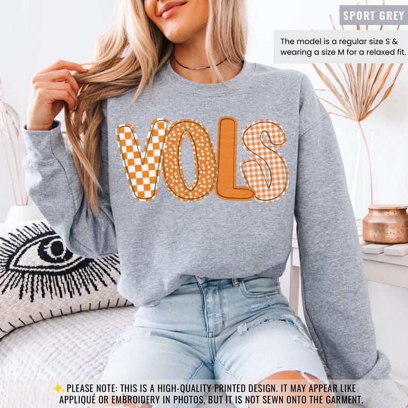 May include: Sport grey sweatshirt with the word "VOLS" in orange and white checkered, polka dot, and gingham patterns. The model is wearing a size M for a relaxed fit. The design is printed on the garment.