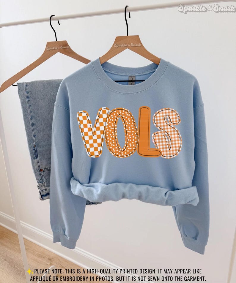 May include: A light blue sweatshirt with the word "VOLS" in orange letters. The letters have a checkerboard and polka dot pattern. The sweatshirt is hanging on a wooden hanger with a pair of blue jeans.