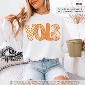 May include: White sweatshirt with the word "VOLS" in orange and patterned letters. The "V" is a checkered pattern, the "O" has polka dots, and the "LS" is a gingham pattern. The model is wearing a size M for a relaxed fit.