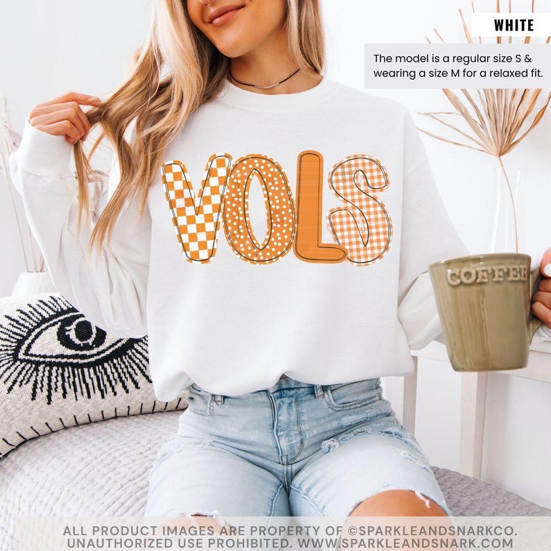 May include: White sweatshirt with the word "VOLS" in orange and patterned letters. The "V" is a checkered pattern, the "O" has polka dots, and the "LS" is a gingham pattern. The model is wearing a size M for a relaxed fit.