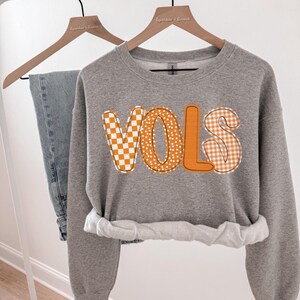 May include: A gray sweatshirt with the word "VOLS" in orange, featuring a checkerboard, polka dot, and gingham pattern. The sweatshirt is hanging on a wooden hanger next to a pair of blue jeans.