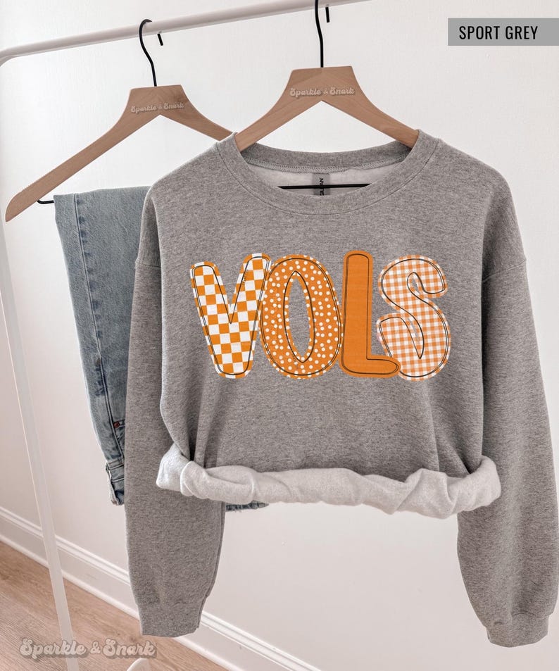 May include: A gray sweatshirt with the word "VOLS" in orange, featuring a checkerboard, polka dot, and gingham pattern. The sweatshirt is hanging on a wooden hanger next to a pair of blue jeans.