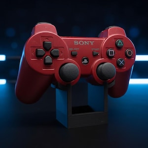 Peut inclure: Une manette de jeu sans fil rouge foncé arborant le logo SONY, les boutons SELECT et START, et un symbole PlayStation central. La manette comprend une croix directionnelle, deux joysticks noirs et quatre boutons d'action avec des icônes triangle, cercle, croix et carré.