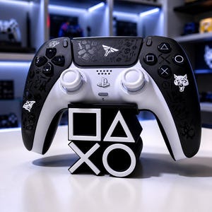 Può includere: Un controller per videogiochi bianco e nero con incisioni personalizzate di simboli e una testa di lupo, poggiato su un supporto bianco e nero a forma di icone dei pulsanti PlayStation: quadrato, triangolo, croce e cerchio.