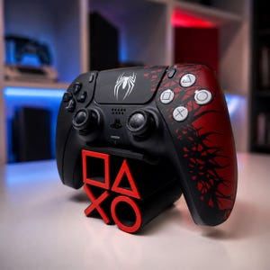 Może przedstawiać: Czarno-czerwony kontroler do gier wideo z białym emblemem pająka na panelu dotykowym oraz czarny stojak z czerwonymi akcentami w kształcie symboli PlayStation: kwadratu, trójkąta, krzyżyka i kółka, spoczywający na płaskiej powierzchni.