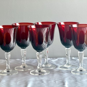 May include: A set of six wine glasses featuring deep red bowls and clear, twisted stems. Each glass has a wide, circular base and a smooth, rounded rim. The bowls are translucent, allowing light to pass through the dark red glass material.