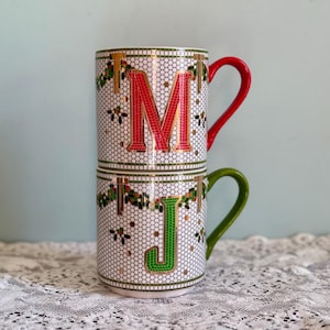 May include: Two stacked ceramic mugs featuring a white honeycomb pattern background with gold accents. The top mug displays a large red letter M and has a red handle. The bottom mug displays a large green letter J and has a green handle.