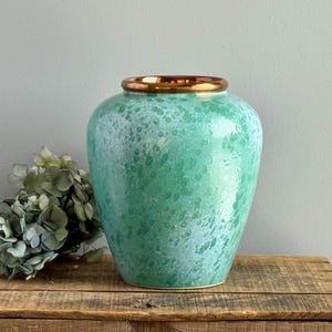 May include: A turquoise ceramic vase with a speckled pattern and a gold rim. The vase has a rounded shape and sits on a wooden surface. Decorative vase for home decor.