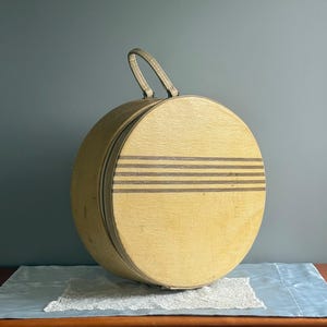 May include: A round, vintage hat box with a tan exterior and three dark gray horizontal stripes. It has a matching handle and is set on a blue cloth with a lace trim. The box is likely made of cardboard or a similar material.